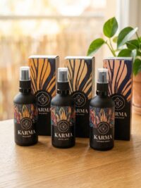 Drei schwarze Sprühflaschen mit bunten, botanischen Etiketten stehen vor passenden Geschenkboxen auf einem hellen Holztisch in Wien Mitte. Die Etiketten tragen die Aufschriften „KARMA WALDBAD“, „KARMA GUTE LAUNE“ und „KARMA INNERER FRIEDE“.